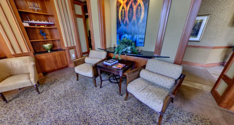 A cozy common-room seating area with upholstered armchairs, a small table with magazines, built-in shelving and wall art.