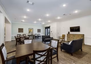 A spacious common area with multiple wooden tables and chairs, two dark upholstered armchairs, a wooden cabinet with a flat-screen TV mounted above it, and framed artwork on the walls. The room has recessed lighting and wood flooring.