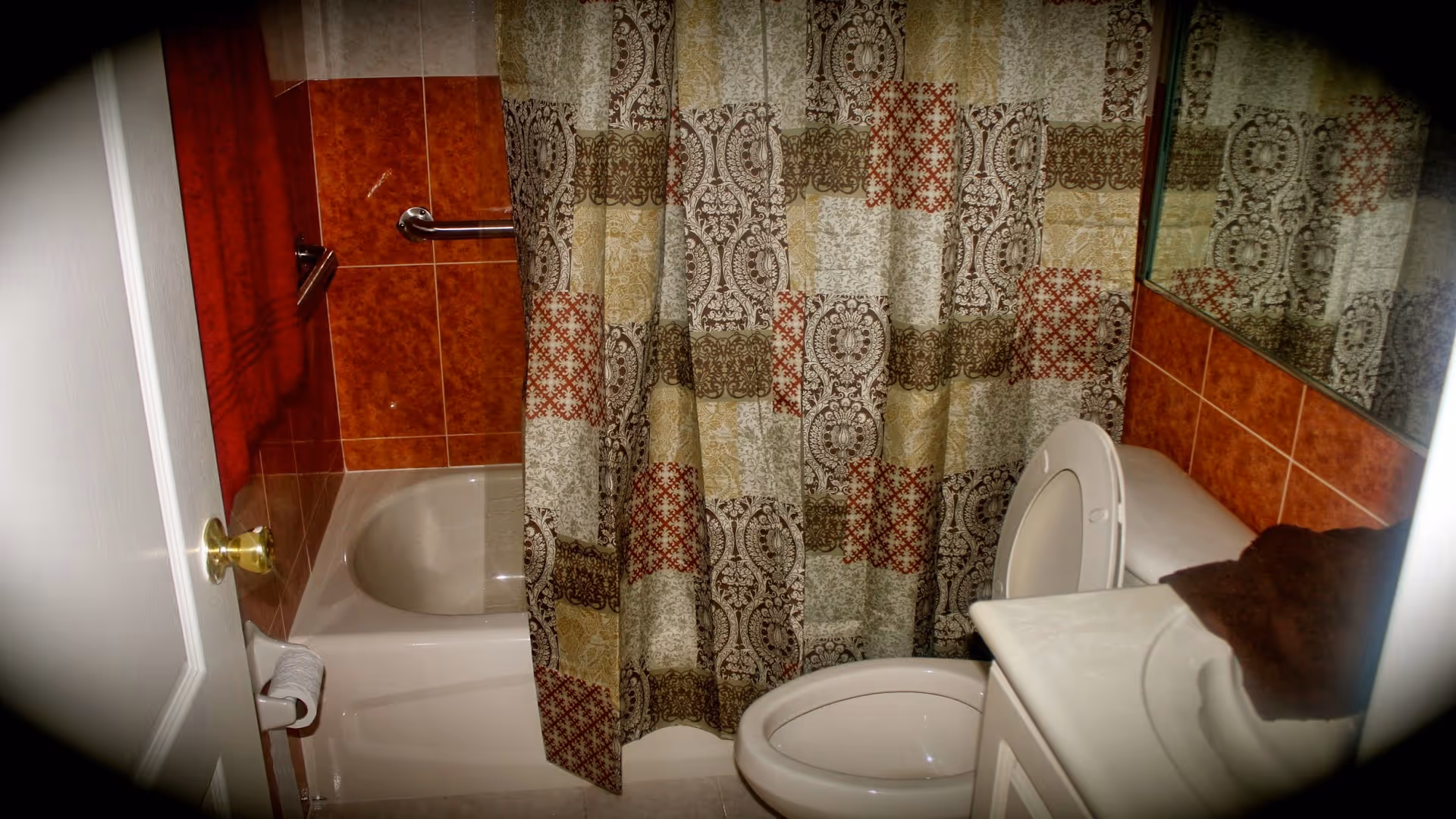A small bathroom with a white bathtub partially covered by a patterned shower curtain in shades of brown, beige, and red. The walls around the bathtub are tiled with reddish-brown tiles. There is a white toilet with the lid open next to a white sink with a brown towel on the counter. A mirror is visible above the sink.