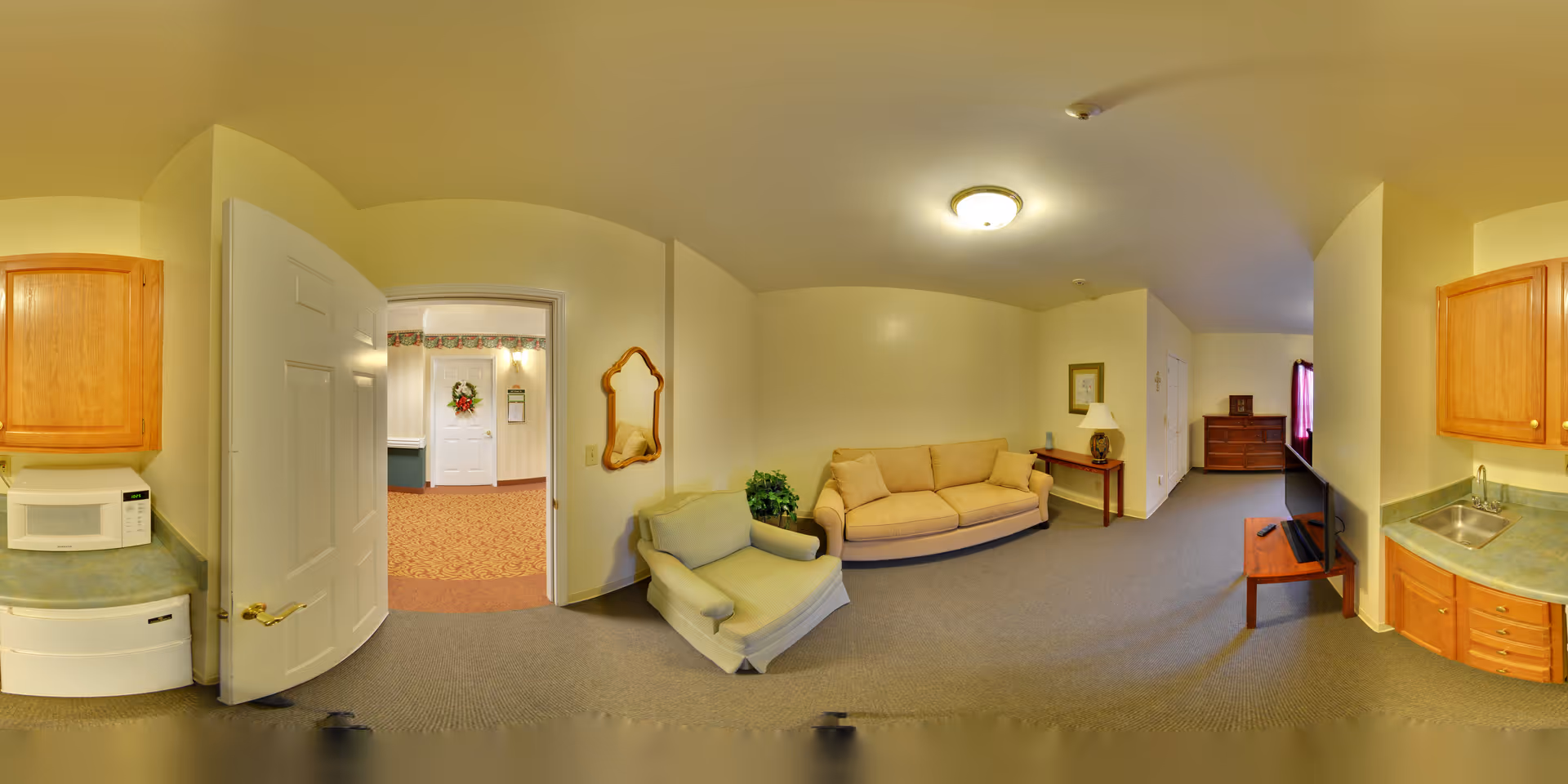 A wide-angle view of a living area in a senior living facility. The room features a beige sofa, a cushioned armchair, a small wooden side table with a lamp, a flat-screen TV on a wooden stand, and a wooden dresser near a window with curtains. The room has light yellow walls and carpeted flooring. On the left side, there is a small kitchenette area with a microwave, dishwasher, sink, and wooden cabinets. An open door leads to a hallway with patterned carpet and a white door decorated with a wreath.