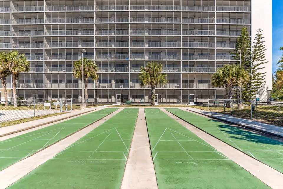 Outdoor shuffleboard courts with green playing surfaces in front of a multi-story residential building with balconies. Palm trees and a chain-link fence surround the area under a clear blue sky.