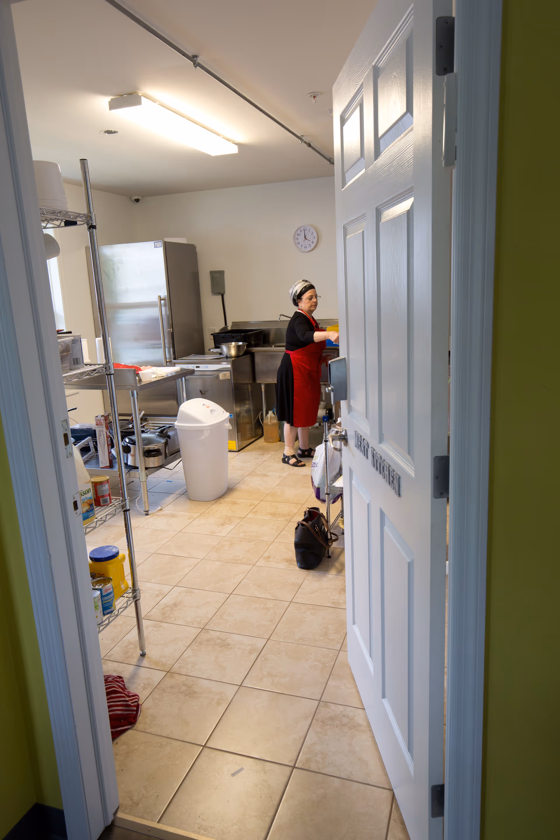 View through an open door into a kitchen where a woman wearing a red apron and headscarf is working at a counter. The kitchen has tiled floors, stainless steel appliances including a refrigerator and sink, metal shelving with various containers, and a white trash bin in the center.