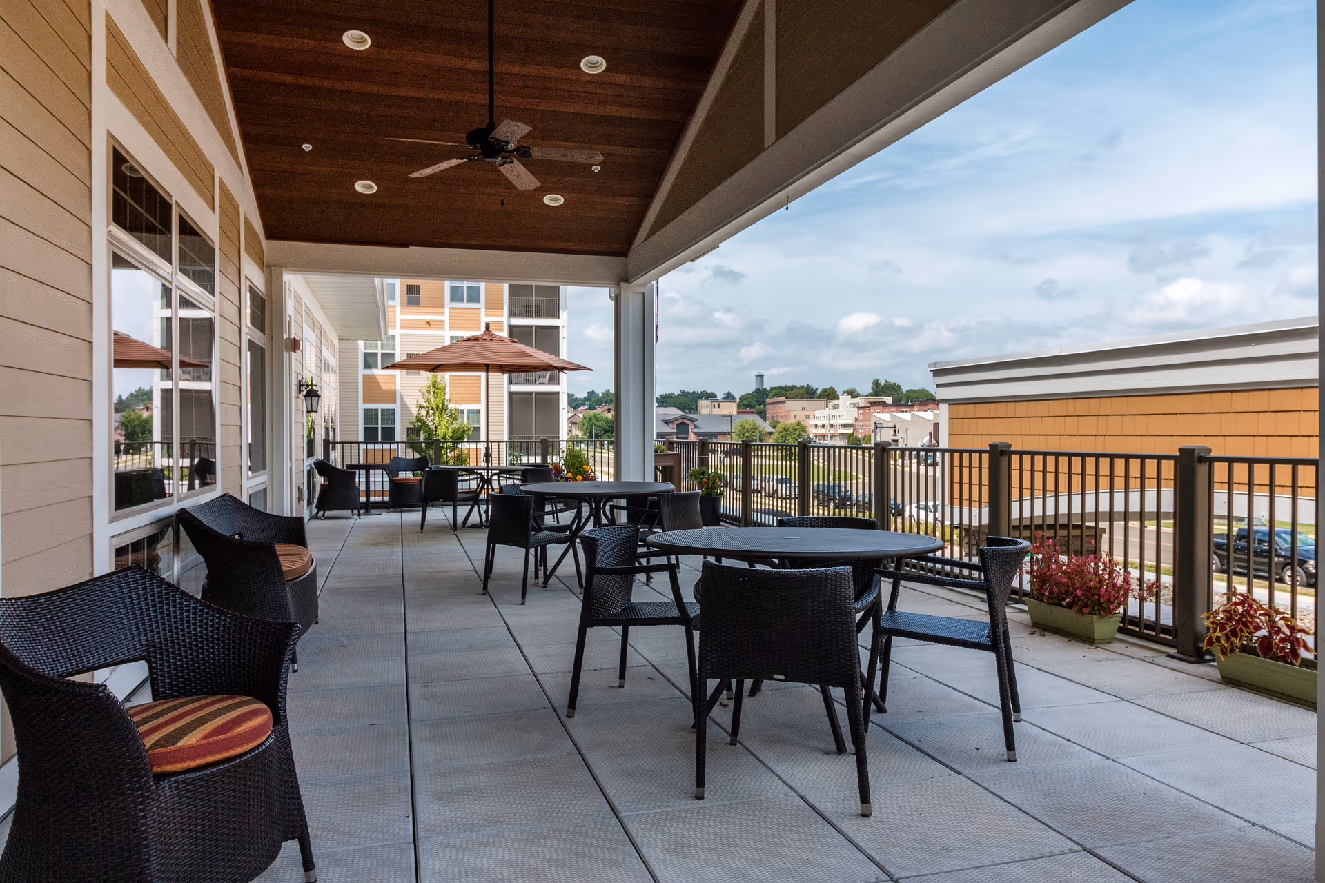 Covered outdoor patio area with multiple black wicker chairs and round tables, some with umbrellas. The patio overlooks a parking lot and nearby buildings under a partly cloudy sky.