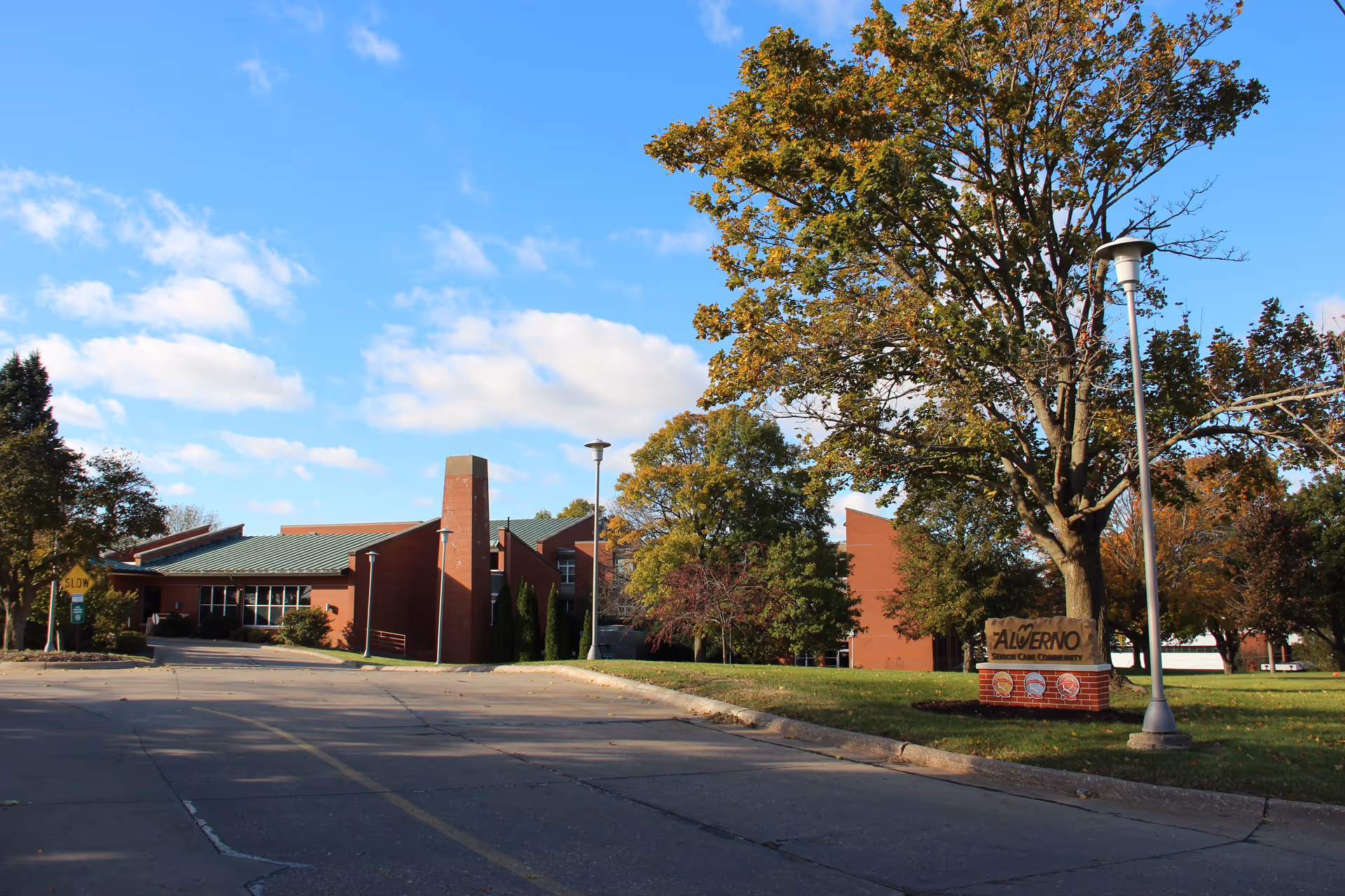 Exterior view of The Alverno Senior Care Community building with a clear blue sky, trees with autumn foliage, a street lamp, and a sign displaying the facility's name on a grassy area near the sidewalk.