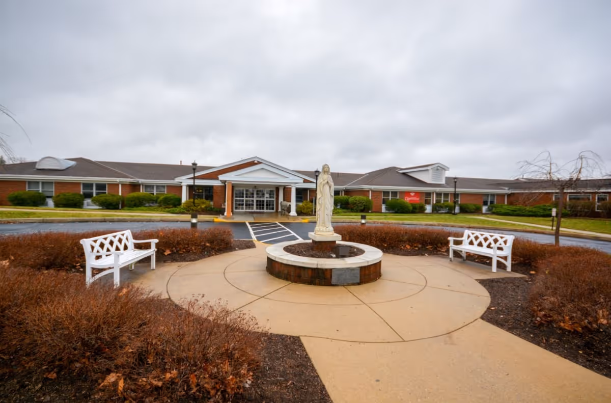Front exterior view of St. Mary Villa for Independent & Retirement Living showing a circular driveway with a statue in the center surrounded by two white benches and landscaped bushes, under a cloudy sky.