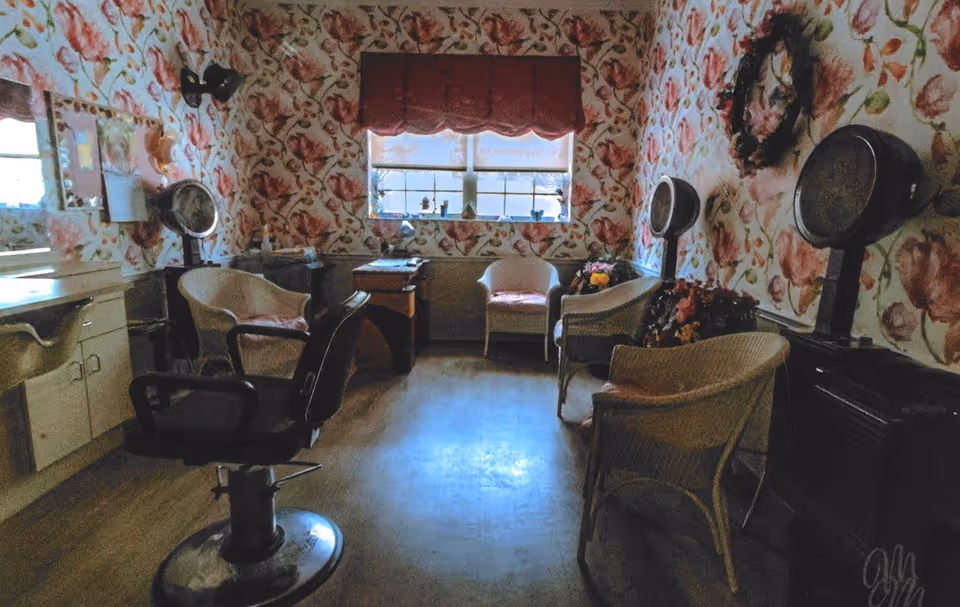 Interior of a vintage-style hair salon with floral wallpaper, several salon chairs including a black styling chair in the foreground, wicker chairs with cushions, hair dryers mounted on stands, a window with a red valance, and a small table with flowers.