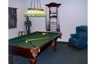 Billiards room with a pool table under a hanging lamp, a cue rack, a blue armchair and a decorative statue.