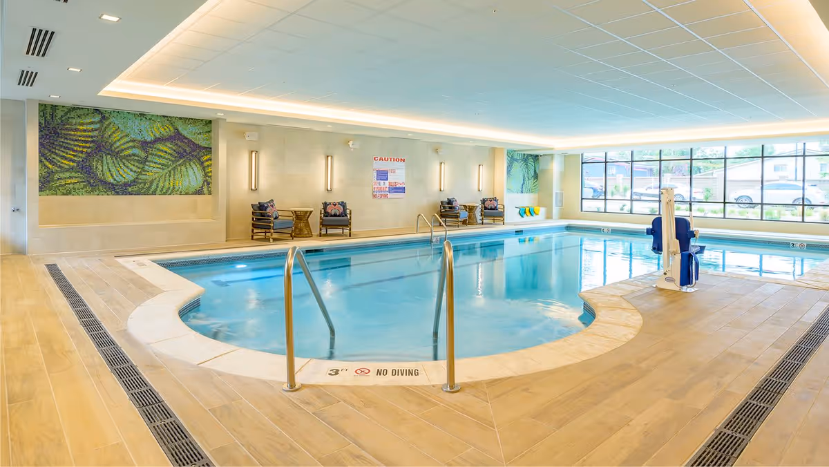 Indoor swimming pool area with clear blue water, metal handrails, pool lift chair, and seating area with chairs and small tables. Large windows allow natural light to fill the space, and decorative wall art is visible on the walls.