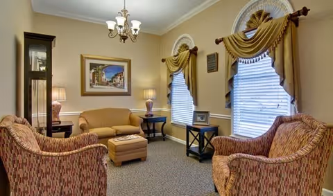 A cozy living room with two patterned armchairs, a beige sofa, and a matching ottoman. The room features two windows with decorative curtains, a grandfather clock, two table lamps on side tables, a chandelier, and a framed painting on the wall.