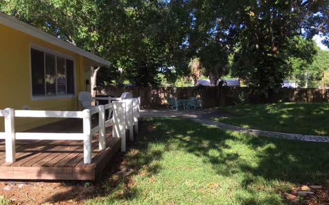 Outdoor area of a facility with a wooden deck attached to a yellow building. The deck has white railings and white chairs. There is a grassy lawn with a stone pathway leading to a seating area with a table and chairs under large trees providing shade.