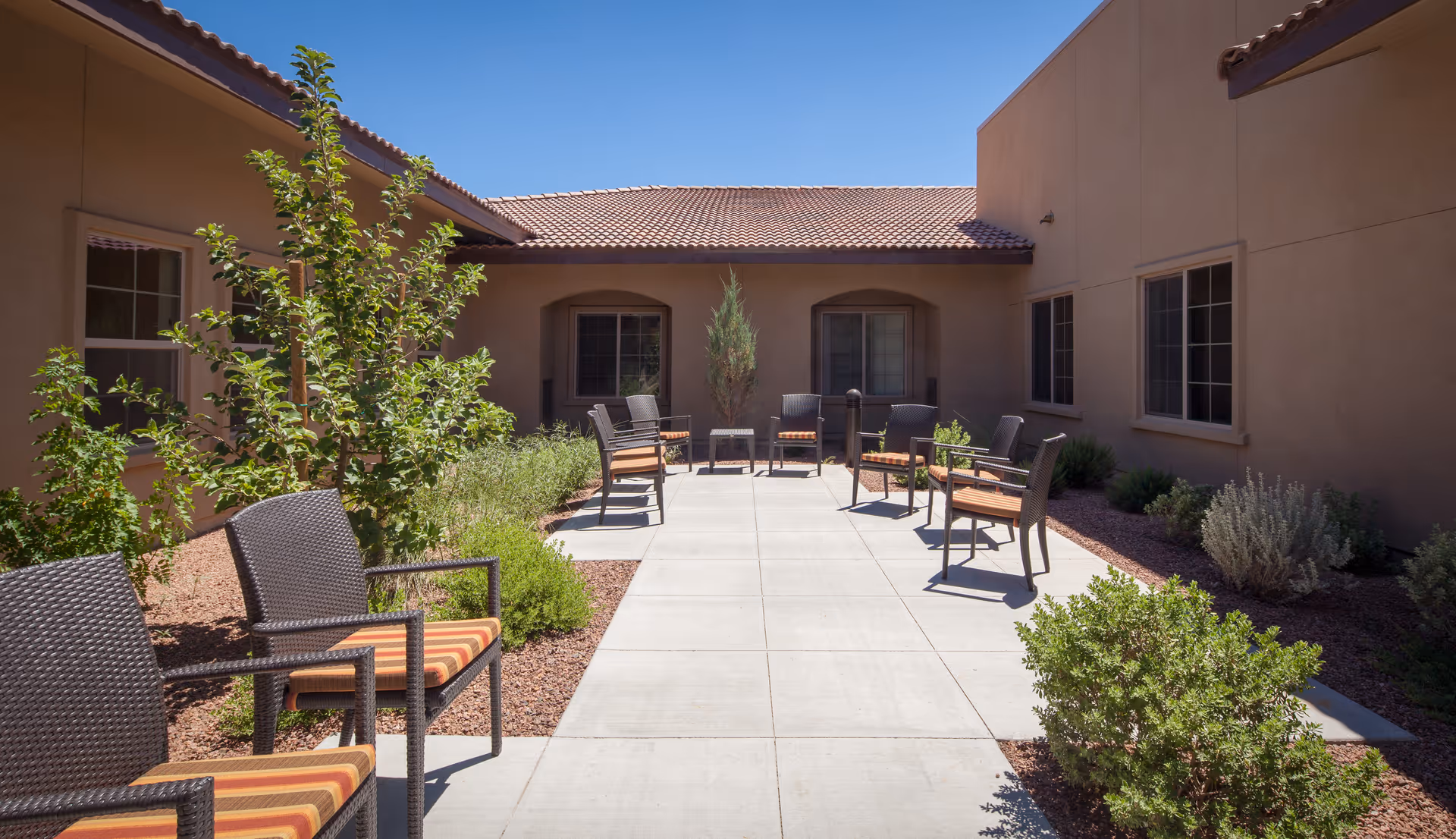 Sunlit courtyard patio with wicker chairs and small tables along a paved walkway between stucco building walls and desert landscaping.