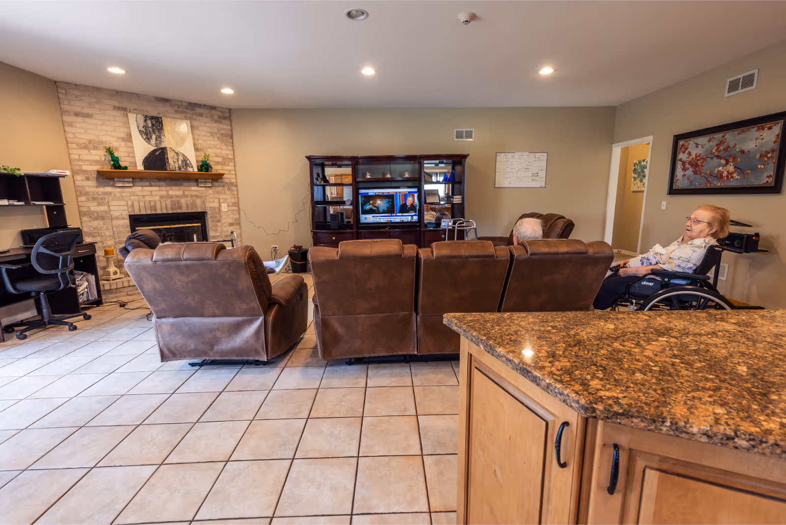 A senior living facility common area with brown leather recliners and a wheelchair. Two elderly individuals are seated facing a television on a wooden entertainment center. The room has tiled flooring, a brick fireplace with a shelf above it, and a desk with an office chair on the left side. The walls are beige with framed artwork and a whiteboard calendar.