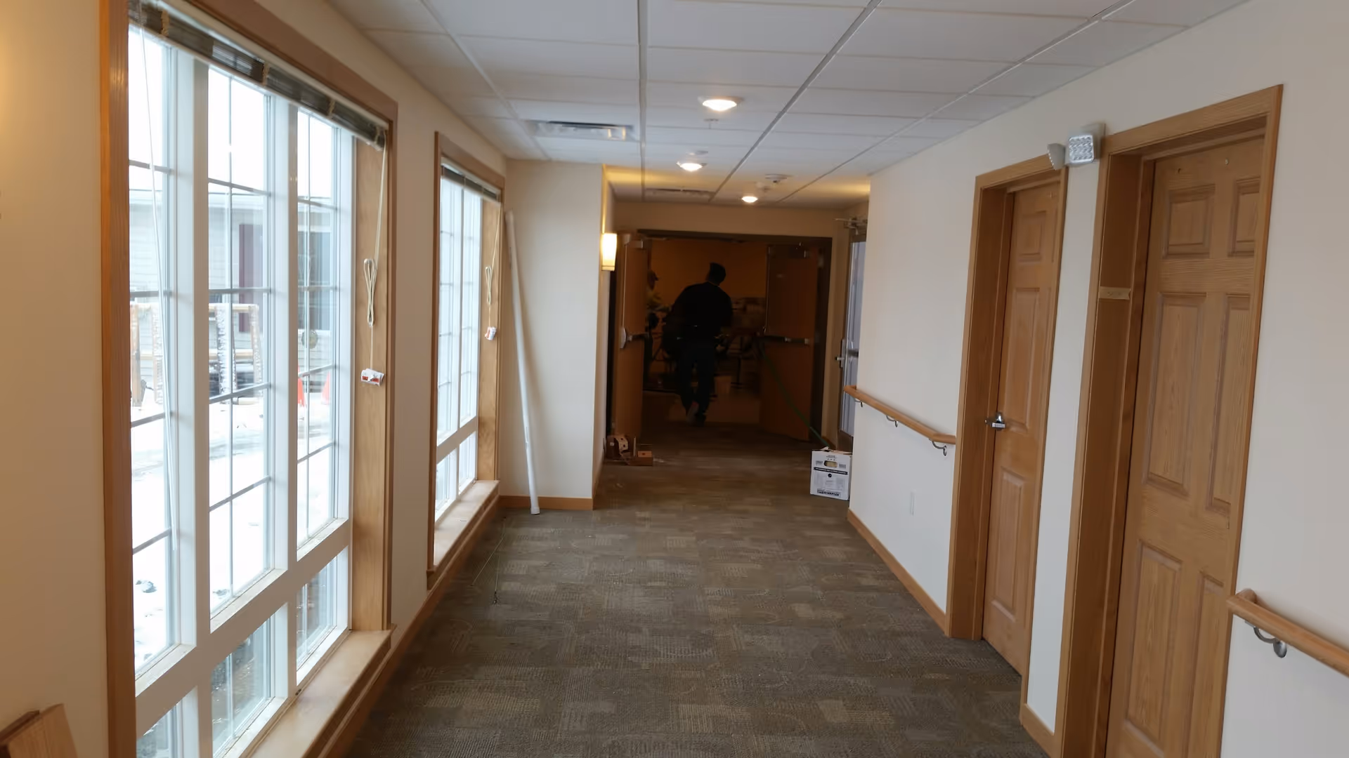 Carpeted interior hallway with large windows on the left, wooden doors and handrails on the right, and a person near the far doorway.