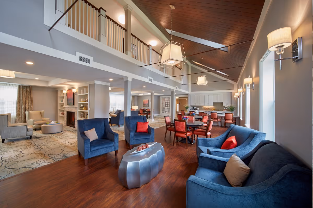 Spacious two-story communal lounge and dining area with blue upholstered seating, dining tables, a fireplace, and a high wood-paneled ceiling.