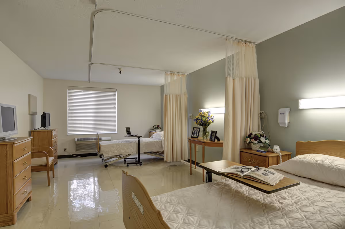 Well-lit shared patient room with two hospital beds separated by privacy curtains, bedside tables, a TV, and an overbed table with a book and glasses.