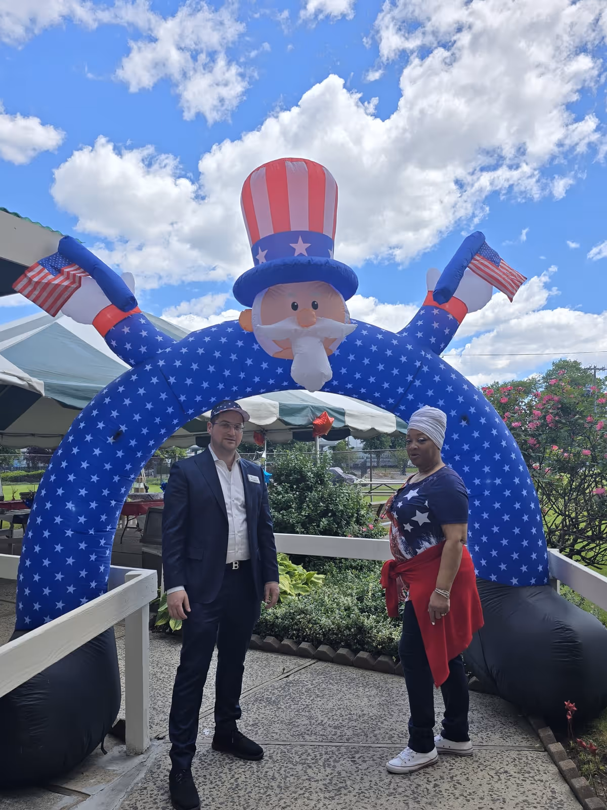 Two people standing outside under a large inflatable arch shaped like Uncle Sam with a red, white, and blue star-spangled design, holding small American flags. The sky is partly cloudy, and there are tents and greenery in the background.