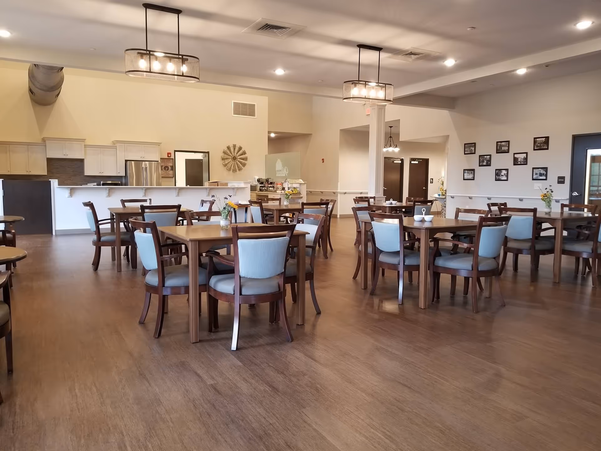 A spacious dining room in an assisted living facility with multiple wooden tables and chairs arranged neatly. The room features modern pendant lighting, a kitchen area with white cabinets and stainless steel appliances in the background, and framed photos on one wall. The floor is wooden, and small flower vases are placed on the tables.