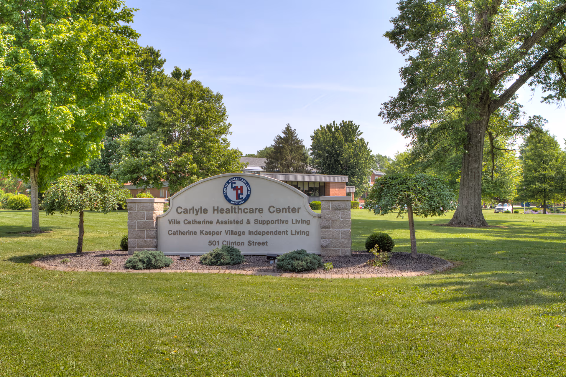 Outdoor view of the Carlyle Healthcare Center sign surrounded by green grass, trees, and shrubs on a sunny day.