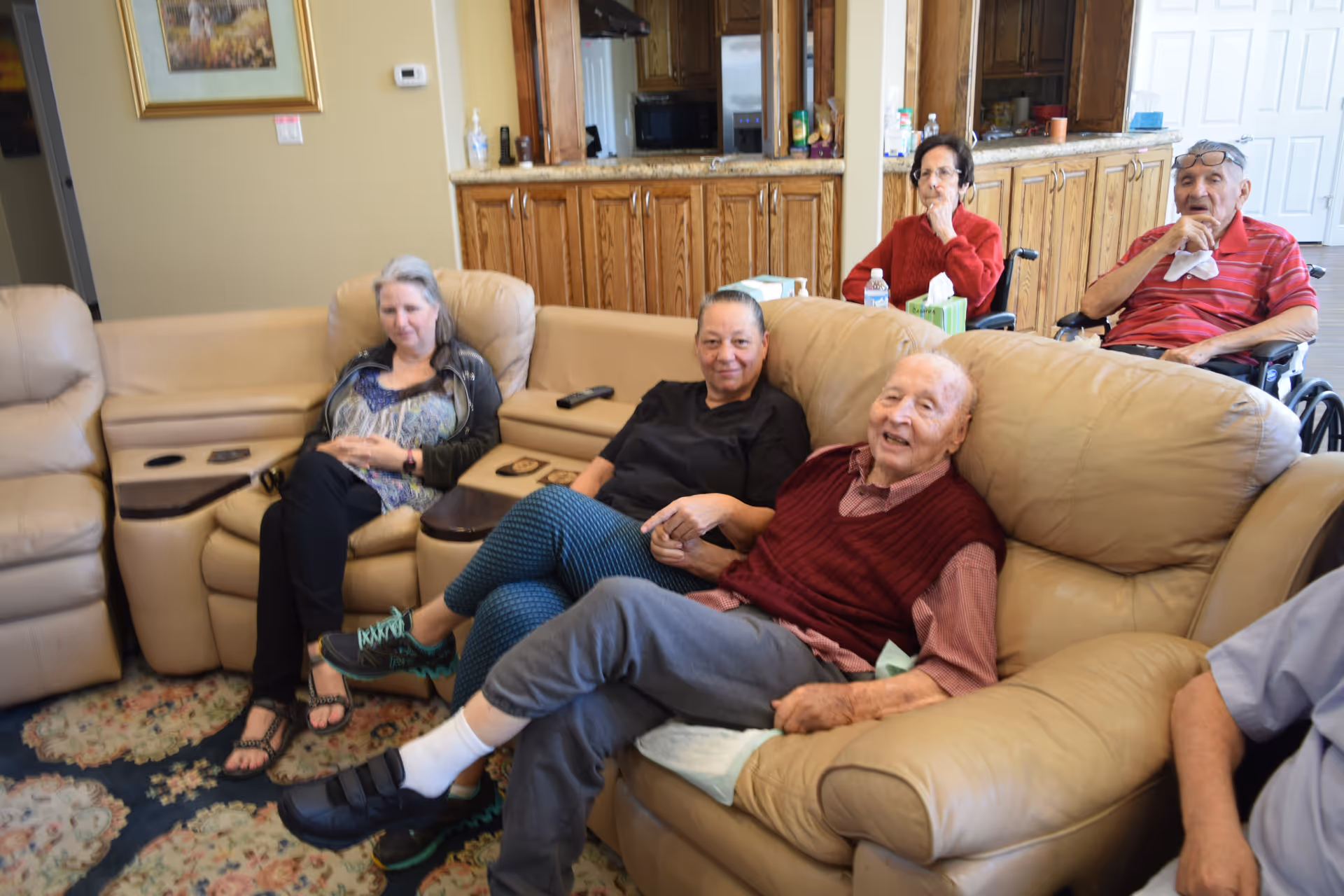 A group of elderly people and a middle-aged woman sitting and relaxing in a living room area with beige leather couches and wooden cabinets in the background. Two elderly individuals are in wheelchairs, and the room has a warm, homey atmosphere.