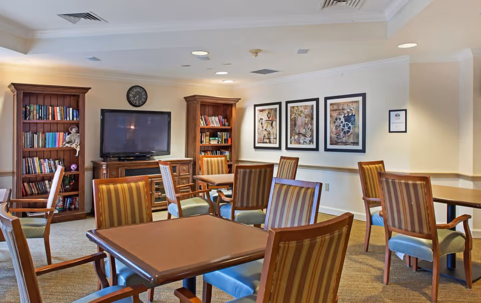 A cozy common area in a senior living facility with several wooden tables and striped cushioned chairs arranged around the room. There are two wooden bookshelves filled with books, a flat-screen TV mounted on a wooden cabinet, and a clock on the wall above the TV. Three framed artworks hang on the wall, and the room is softly lit with ceiling lights.
