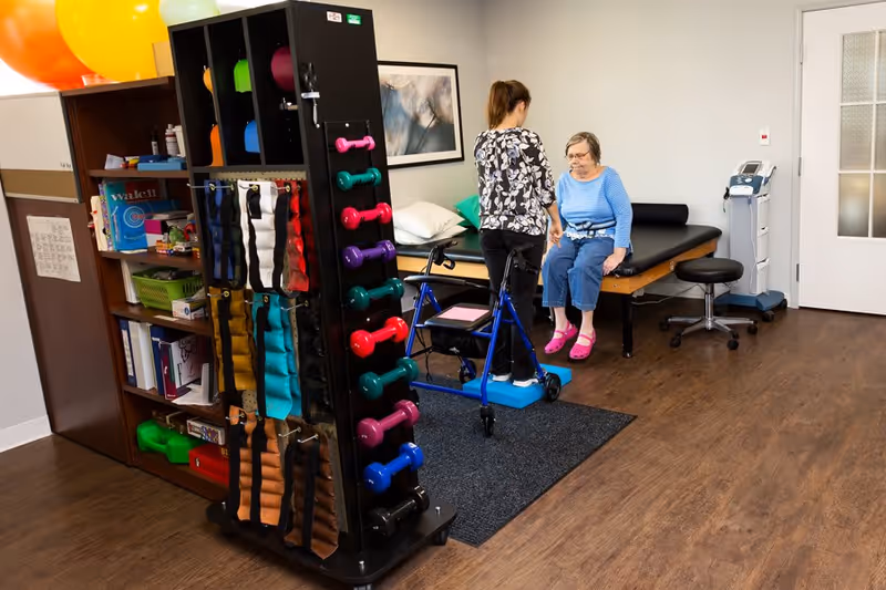 A therapy room in a senior living facility with a therapist assisting an elderly woman sitting on a treatment table. The room contains exercise equipment including colorful dumbbells, resistance bands, large exercise balls, and a walker. The floor is wooden, and there is a medical device near a closed door in the background.