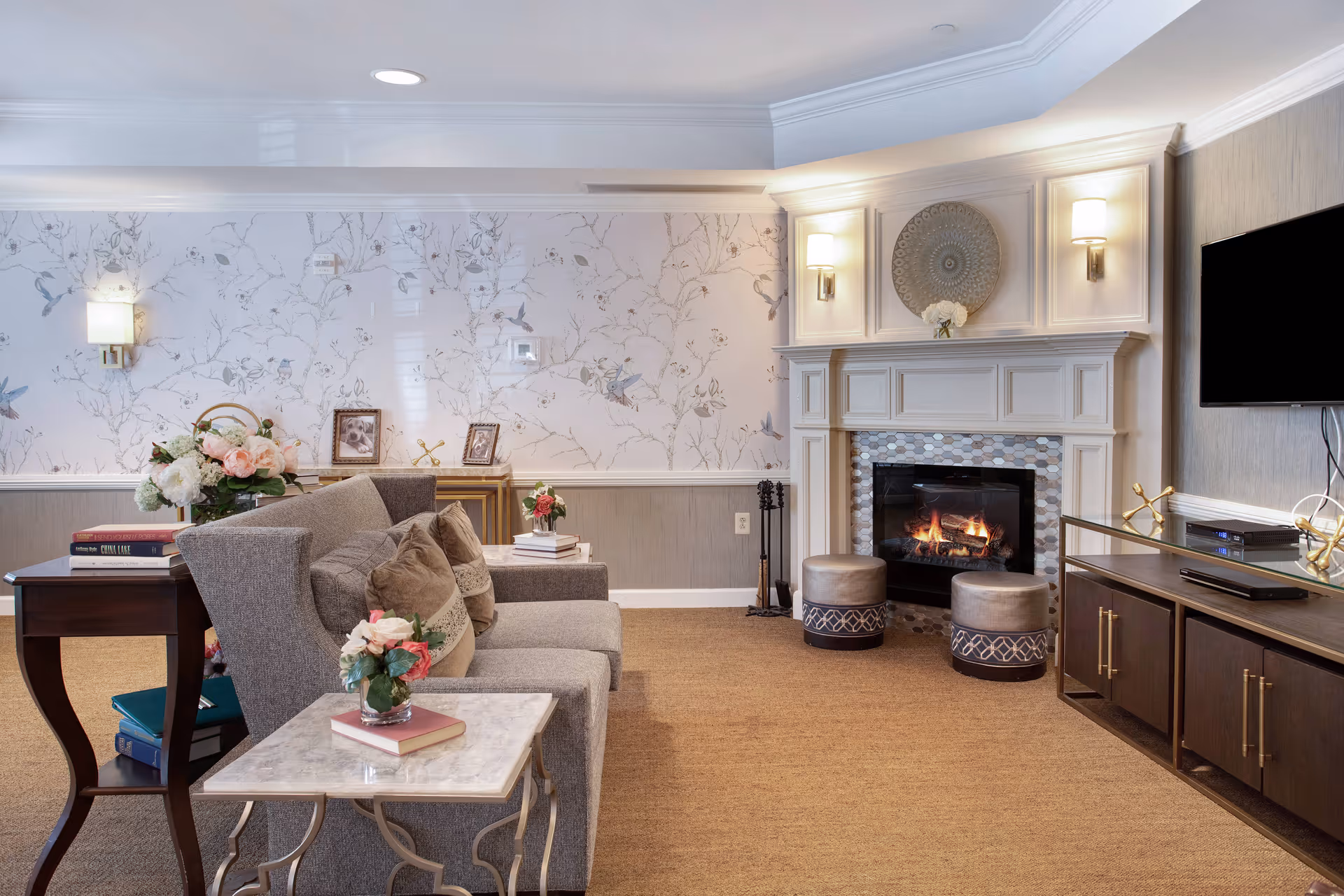 A cozy living room with a gray sofa adorned with pillows, a marble-top side table with a small flower arrangement and book, a wooden side table with books and flowers, a fireplace with a decorative mantle and two round ottomans in front, a wall-mounted flat screen TV, and floral wallpaper on the walls.