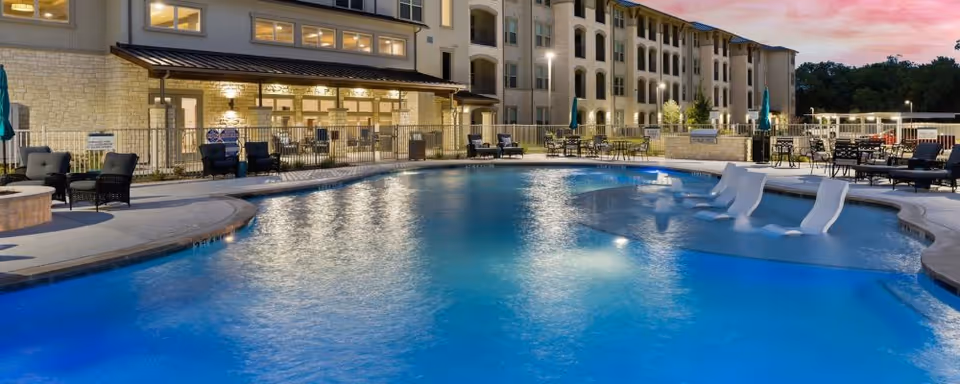 Outdoor swimming pool with built-in lounge seats, patio furniture, and a multi-story building lit at dusk.