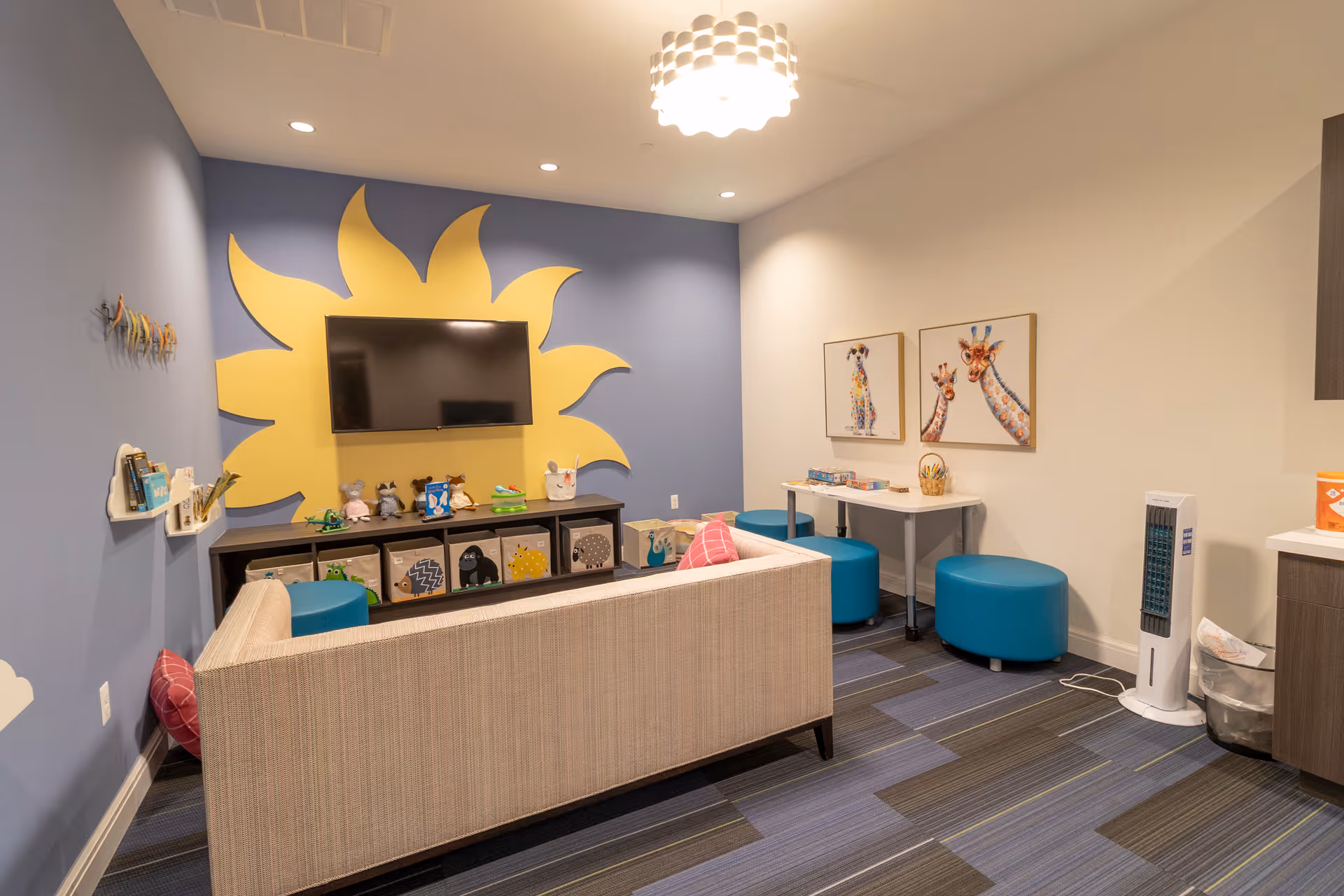 A cozy room with a beige couch facing a wall-mounted TV on a blue wall with a large yellow sun design. Below the TV is a shelf with various toys and storage bins. To the right, there is a small table with board games and two blue round stools. The room has colorful carpet tiles and two framed pictures of giraffes on the wall.