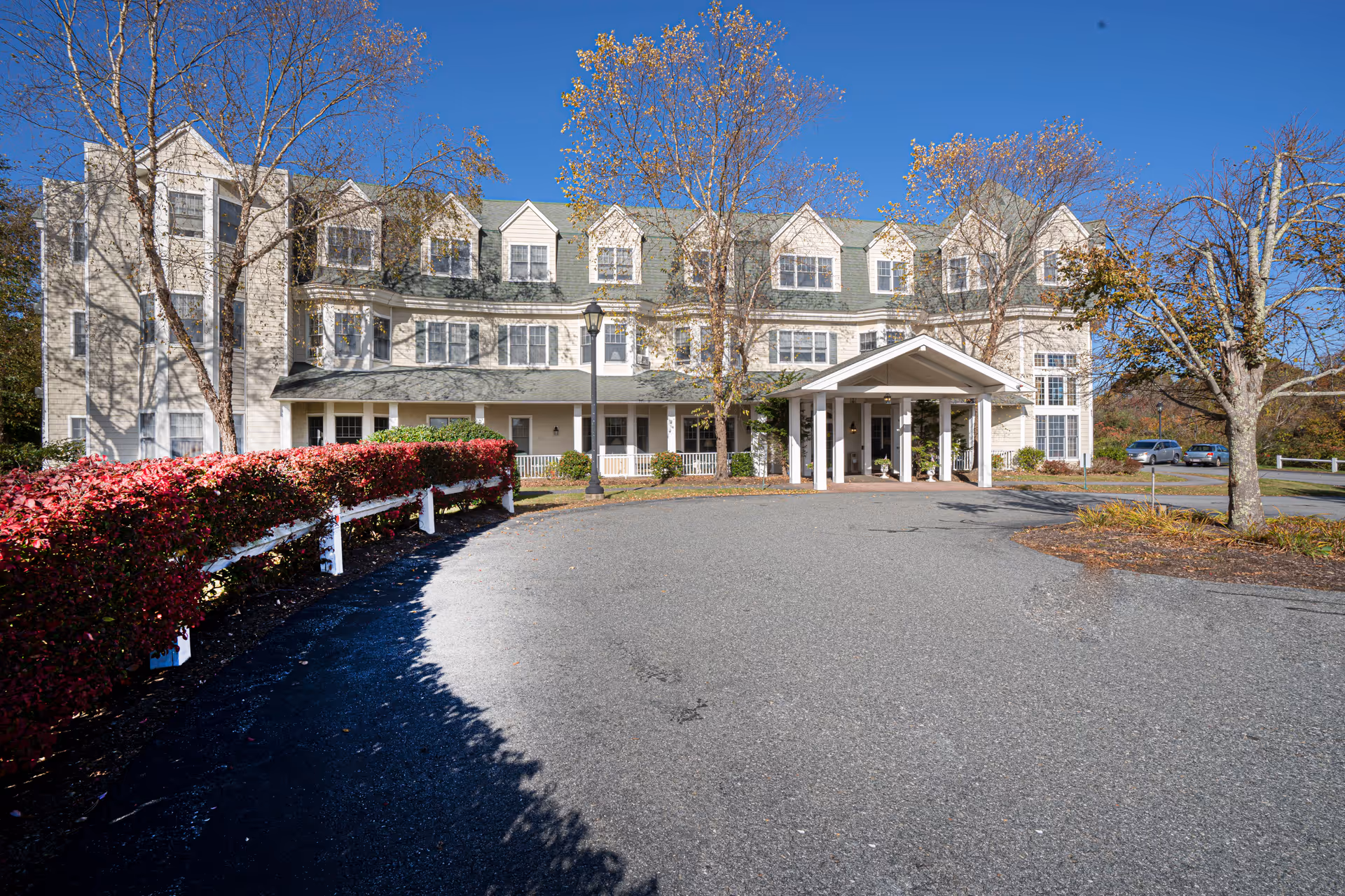 Front exterior view of a three-story assisted living community building with a covered entrance, surrounded by trees and bushes under a clear blue sky.