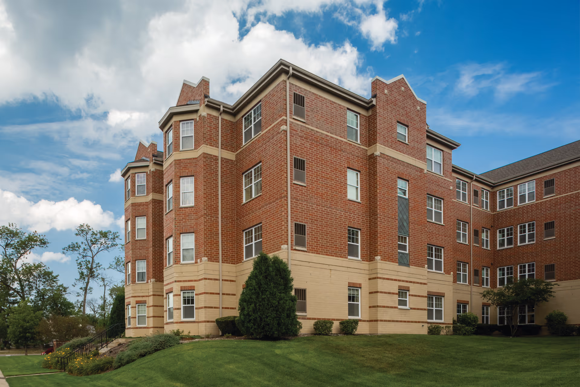 Exterior view of a multi-story brick building with beige accents, surrounded by a well-maintained lawn and some trees under a partly cloudy blue sky.