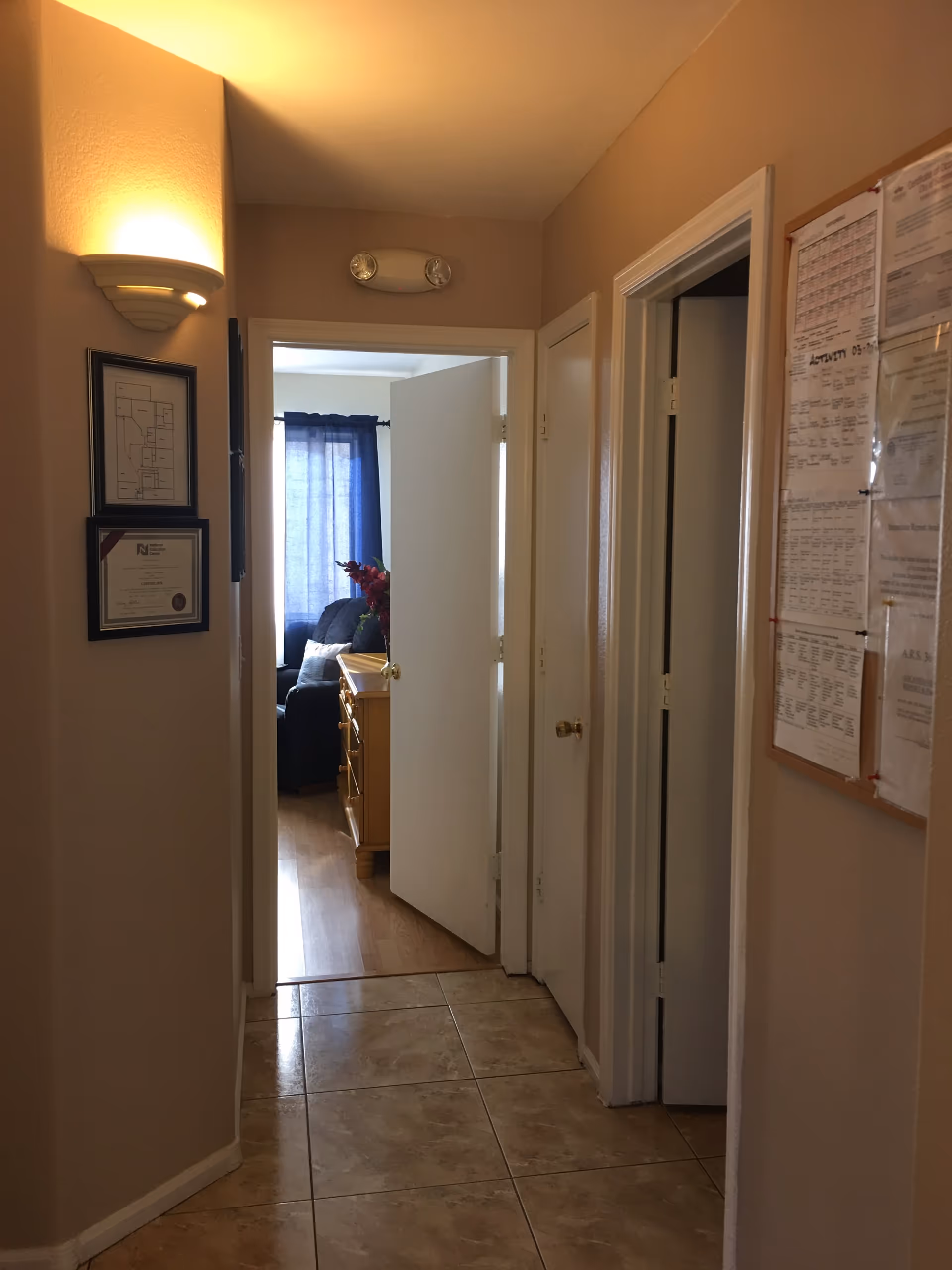 A hallway with beige walls and tiled floor leading to a room with a wooden dresser and a dark armchair near a window with blue curtains. On the left wall of the hallway, there are two framed items, one appears to be a floor plan and the other a certificate. On the right wall, there is a bulletin board with various papers pinned to it. The hallway has three doors, one open leading to the room and two closed.