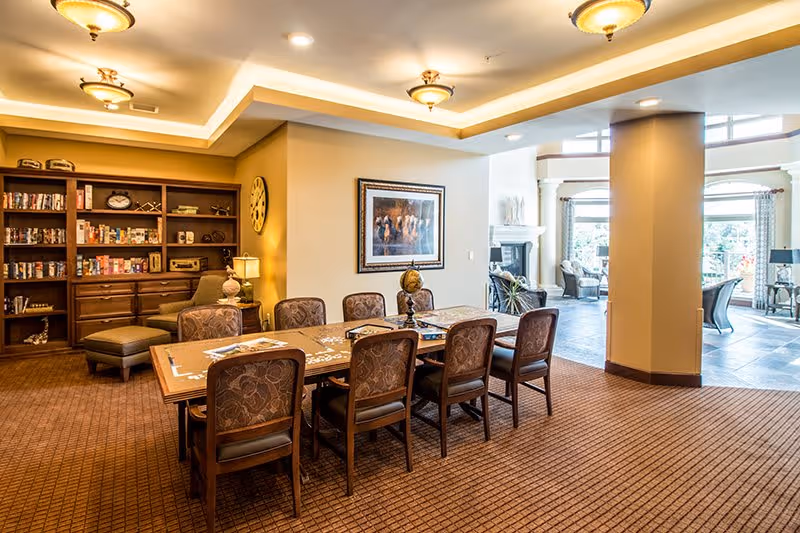 A well-lit common area in a senior living facility featuring a large wooden table with eight upholstered chairs arranged around it. The table has puzzles and games on it. Behind the table is a wooden bookshelf filled with books, games, and decorative items. A comfortable armchair with an ottoman is positioned near the bookshelf. The room has warm lighting with ceiling fixtures and a framed picture on the wall. In the background, there is an open space with large windows letting in natural light and additional seating areas.