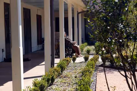 Resident in a wheelchair sits along a shaded walkway beside a columned building