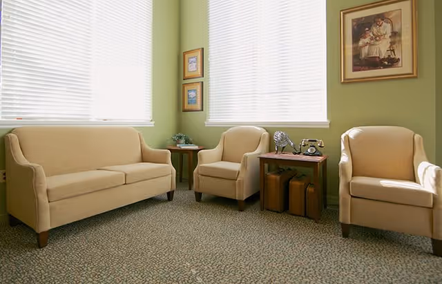 A cozy sitting area with a beige sofa and two matching armchairs arranged around two small wooden side tables. The walls are painted light green and decorated with framed artwork. Two windows with white blinds let in natural light.