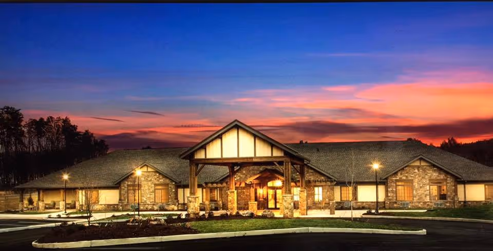 Exterior view of a single-story senior living facility building with stone and wood accents under a colorful sunset sky. The entrance is well-lit with a covered porch supported by stone pillars, surrounded by landscaped greenery and a paved driveway.