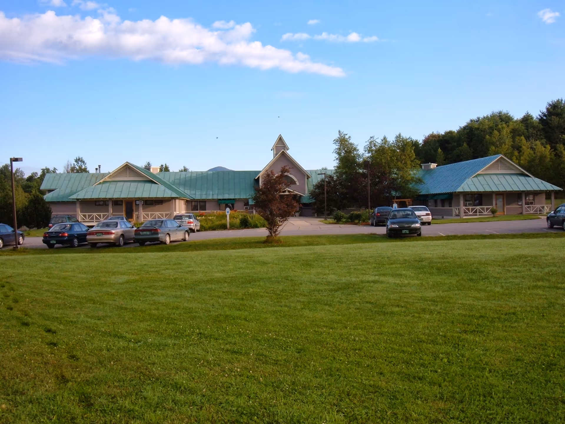Exterior view of a single-story building with a green metal roof and beige walls, surrounded by a grassy lawn and several parked cars under a blue sky with some clouds.