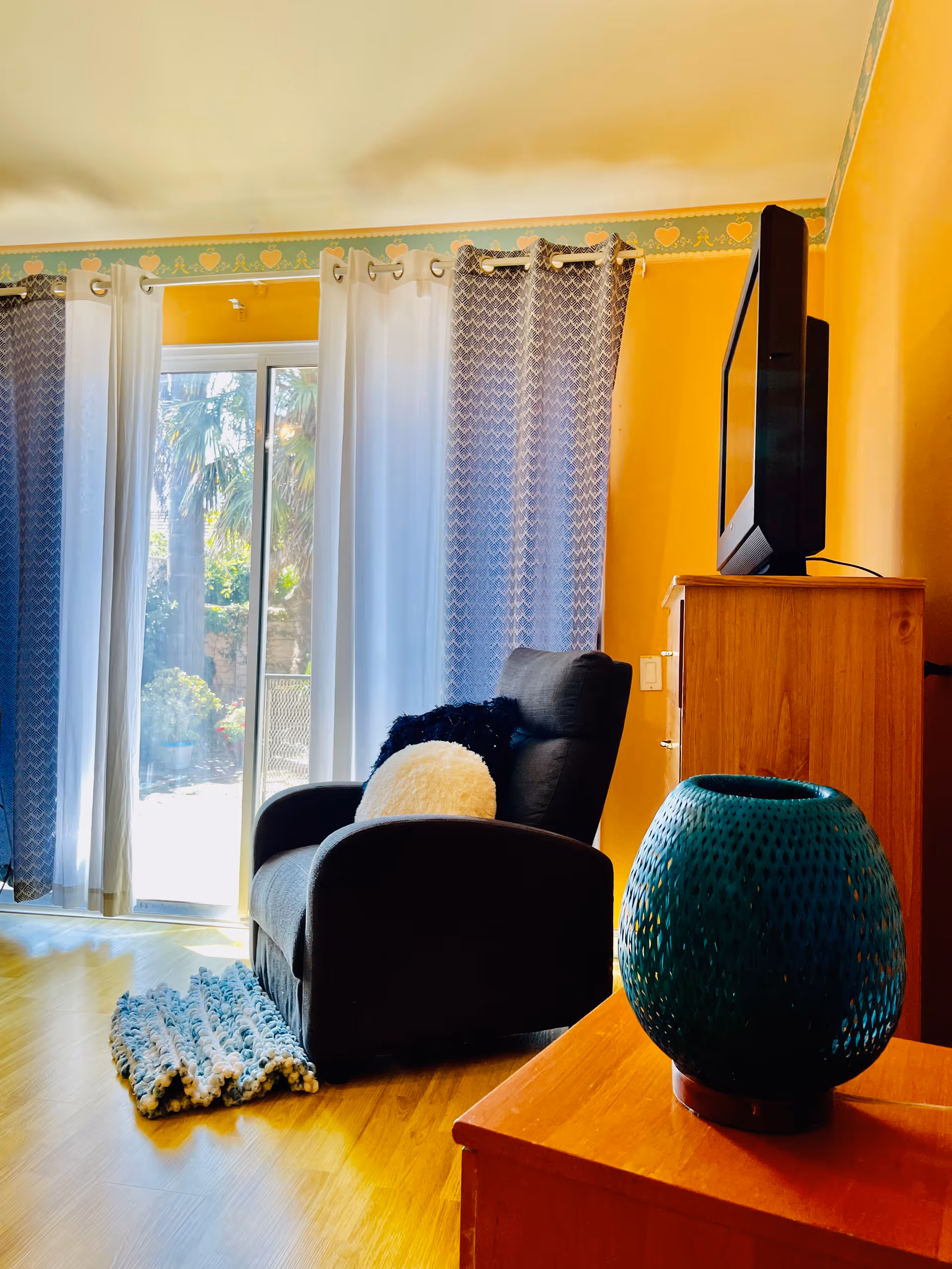 A cozy living room area with a black armchair adorned with two fluffy pillows, one black and one white. The room has yellow walls and a wooden floor. There are sheer white and patterned blue curtains hanging in front of a sliding glass door that leads outside to a garden with palm trees. A wooden dresser with a flat-screen TV on top is visible, along with a blue decorative vase on a wooden surface in the foreground.