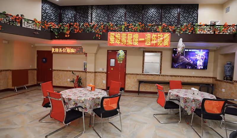 Interior view of a decorated common area with round tables covered in floral tablecloths and surrounded by red and black chairs. There are balloons tied to some chairs, a TV mounted on the wall displaying a scene, and a red banner with yellow Chinese characters hanging above two red doors. The upper wall features a decorative black lattice with green and orange garlands.