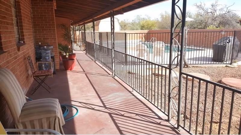 Covered outdoor patio area with a metal railing overlooking a fenced swimming pool. The patio has a few chairs, a potted plant, and a water hose coiled on the floor. The area is adjacent to a brick building wall.