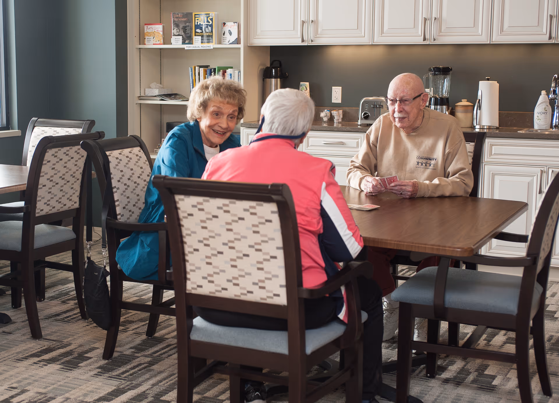 Three elderly people sitting around a table in a well-lit room with kitchen cabinets in the background, playing cards and engaging in conversation.