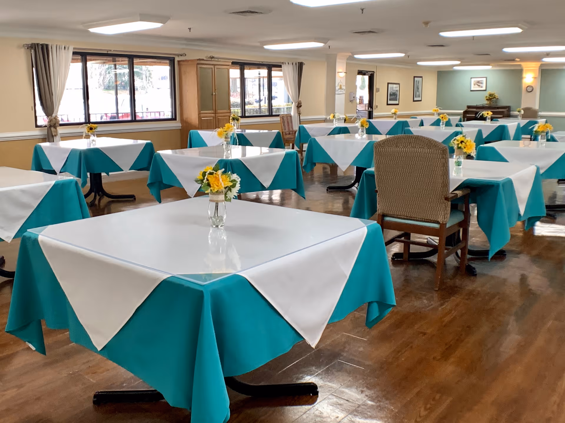 Spacious dining room with multiple tables covered in teal and white tablecloths, each topped with small vases of yellow flowers.