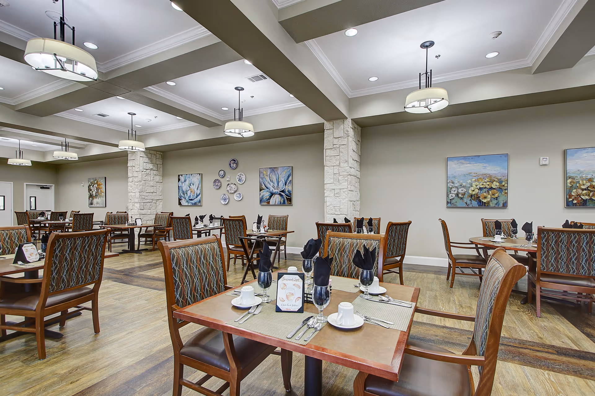 A spacious dining room with multiple wooden tables and chairs arranged neatly. Each table is set with white cups, saucers, silverware, and black cloth napkins in glasses. The room features light-colored walls adorned with floral paintings and decorative plates. The ceiling has exposed beams with modern hanging light fixtures, and the floor is a wood pattern.