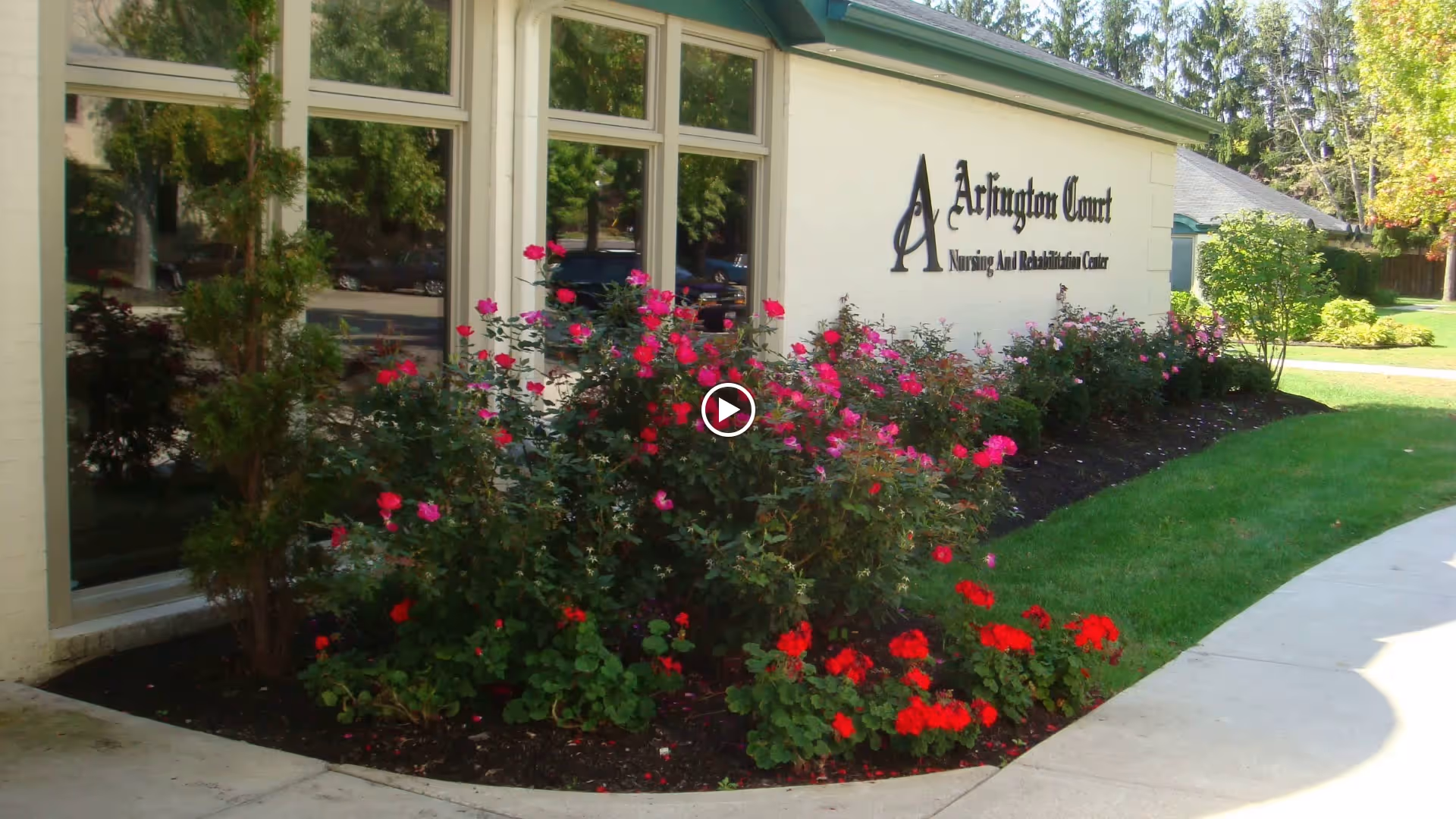 Exterior view of Arlington Court Nursing and Rehabilitation Center building with large windows and a garden bed filled with blooming red and pink flowers along the sidewalk.