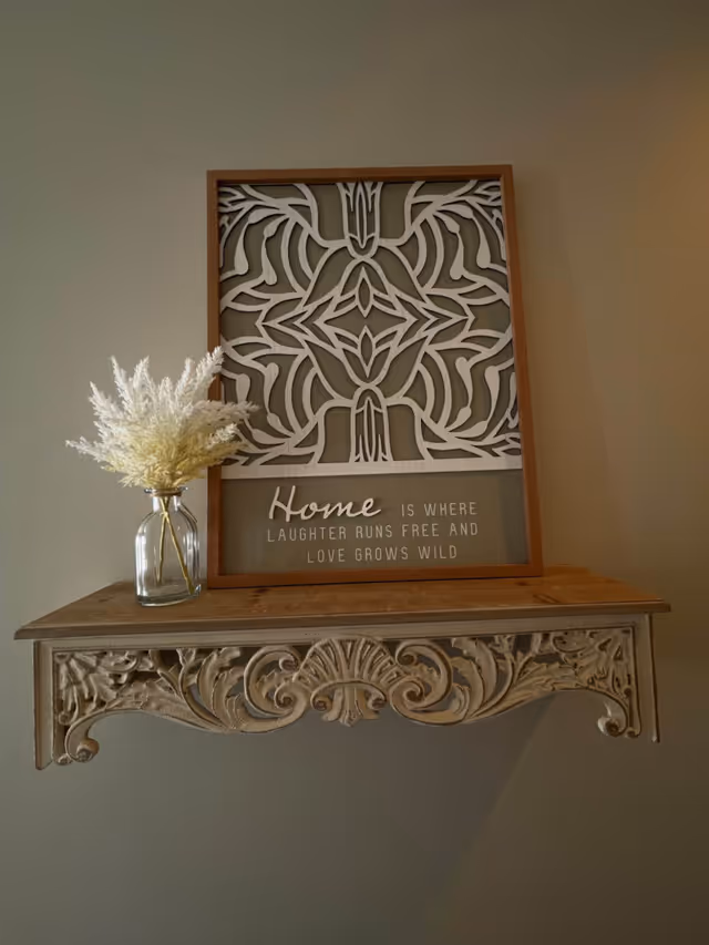 Ornate wooden wall shelf holding a framed decorative 'Home' sign and a glass vase of dried flowers against a beige wall.