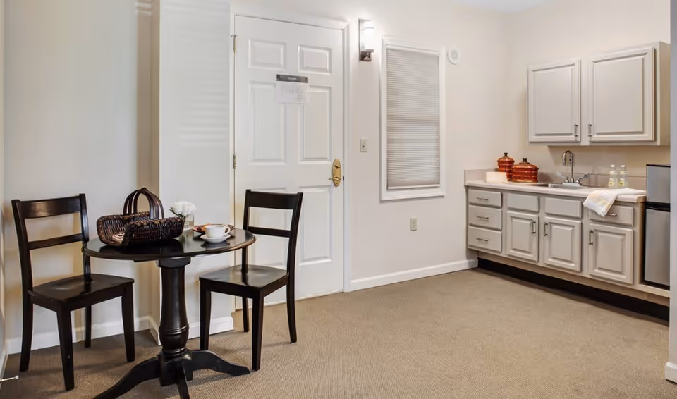 Small dining area with a round black table and two black chairs next to a white door. The table has a woven basket, a cup and saucer, and a small flower vase. To the right, there is a kitchenette with white cabinets, a sink, countertop, and a mini refrigerator. The walls are white and the floor is carpeted.