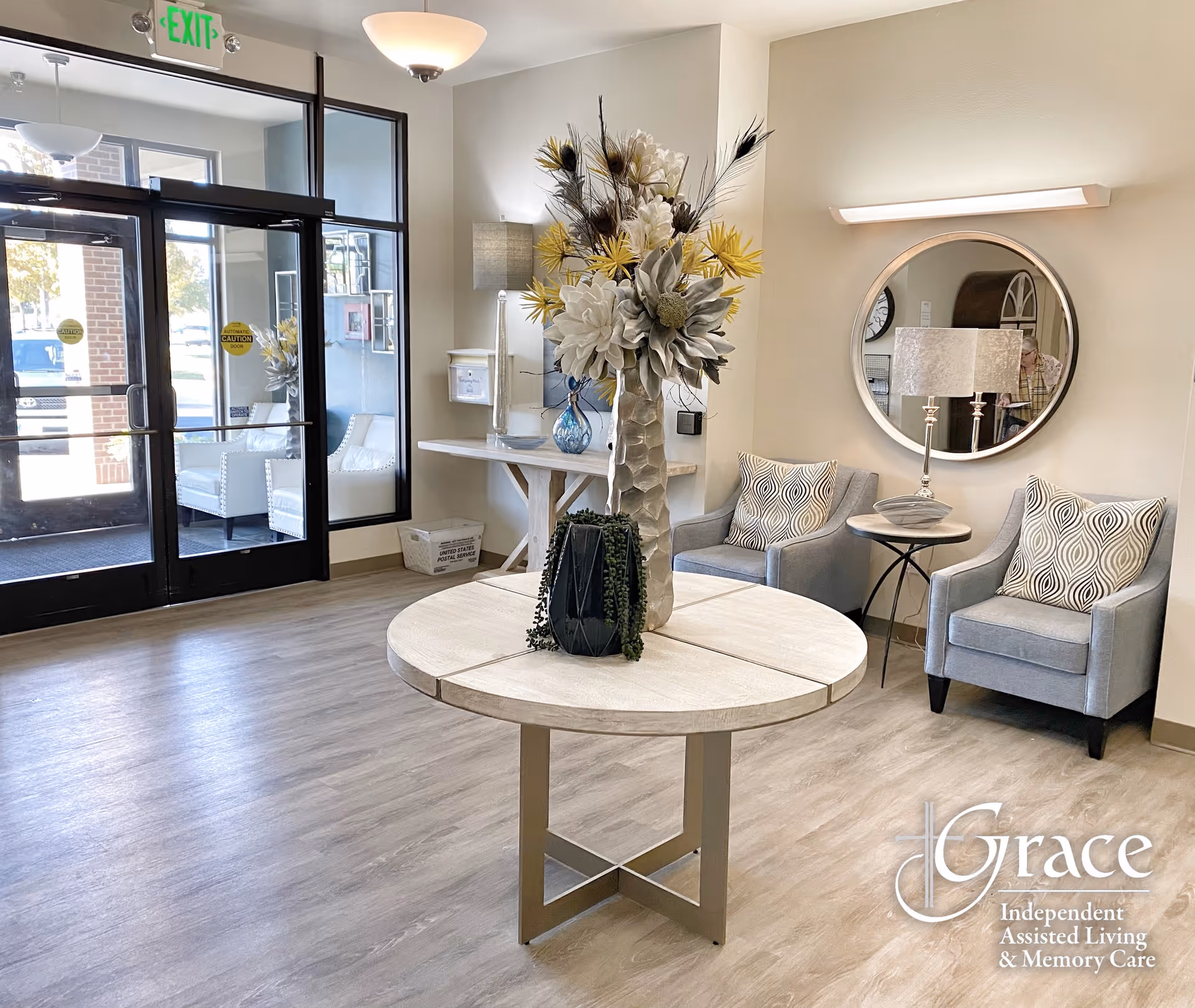 Bright assisted living lobby with a round centerpiece table, floral arrangement, seating area, and glass entry doors.