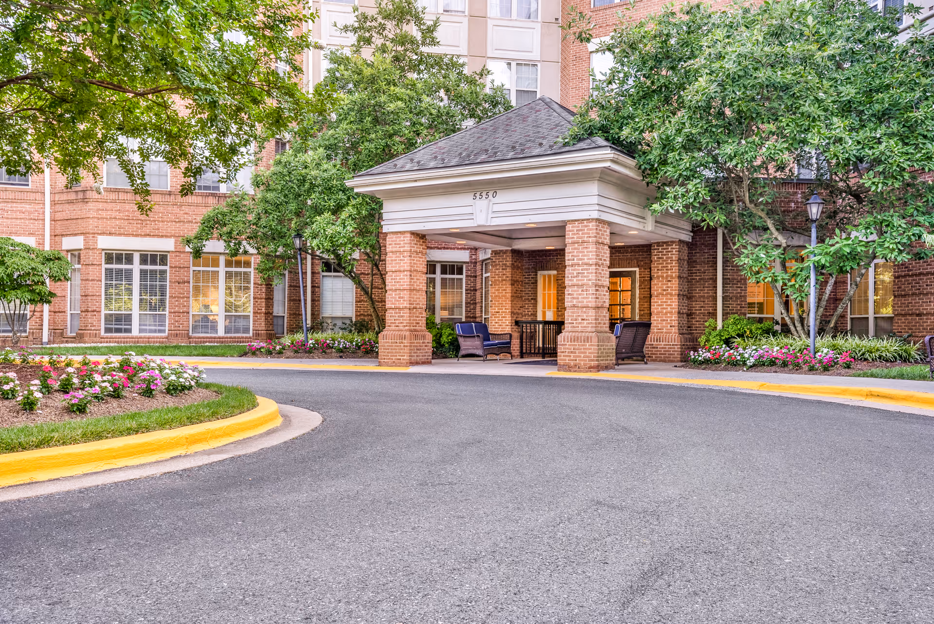 Entrance to a senior living facility with a covered drop-off area supported by brick columns. There are outdoor chairs and a table under the covered area. The building is made of red brick with large windows, surrounded by green trees and colorful flower beds along the driveway.