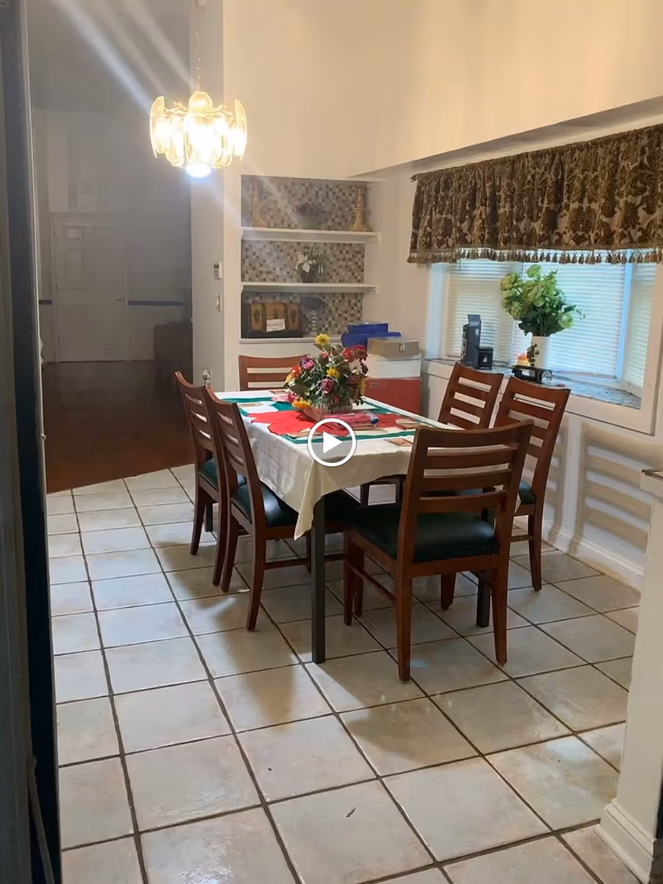 Dining room with a rectangular table covered by a tablecloth and floral centerpiece, six wooden chairs, tiled floor, and a window with a valance.