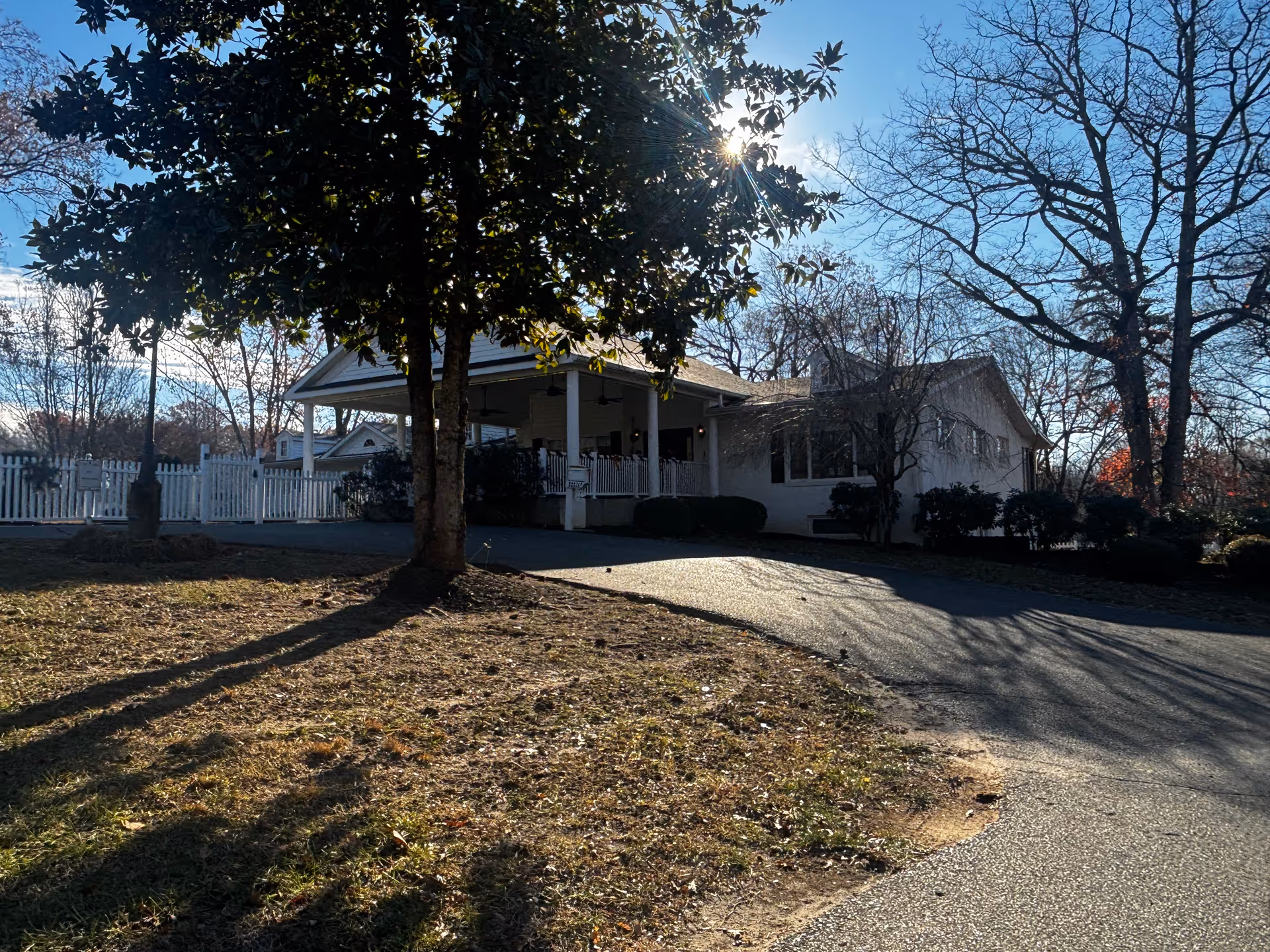 Exterior view of a single-story building with a covered porch, surrounded by trees and bushes. The sun is shining through the leaves of a large tree in the foreground, casting shadows on the ground and driveway. A white picket fence is visible in the background.