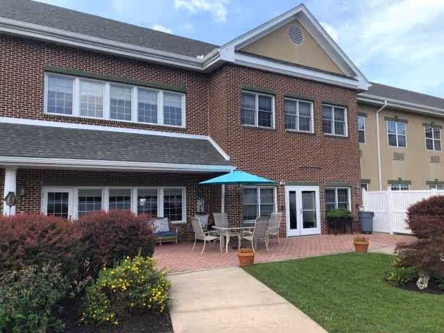 Exterior view of a two-story brick and beige building with multiple windows. There is a patio area with outdoor furniture including a table, chairs, and a blue umbrella. The patio is surrounded by potted plants and landscaping with bushes and flowers. A concrete walkway leads to the patio from the foreground.